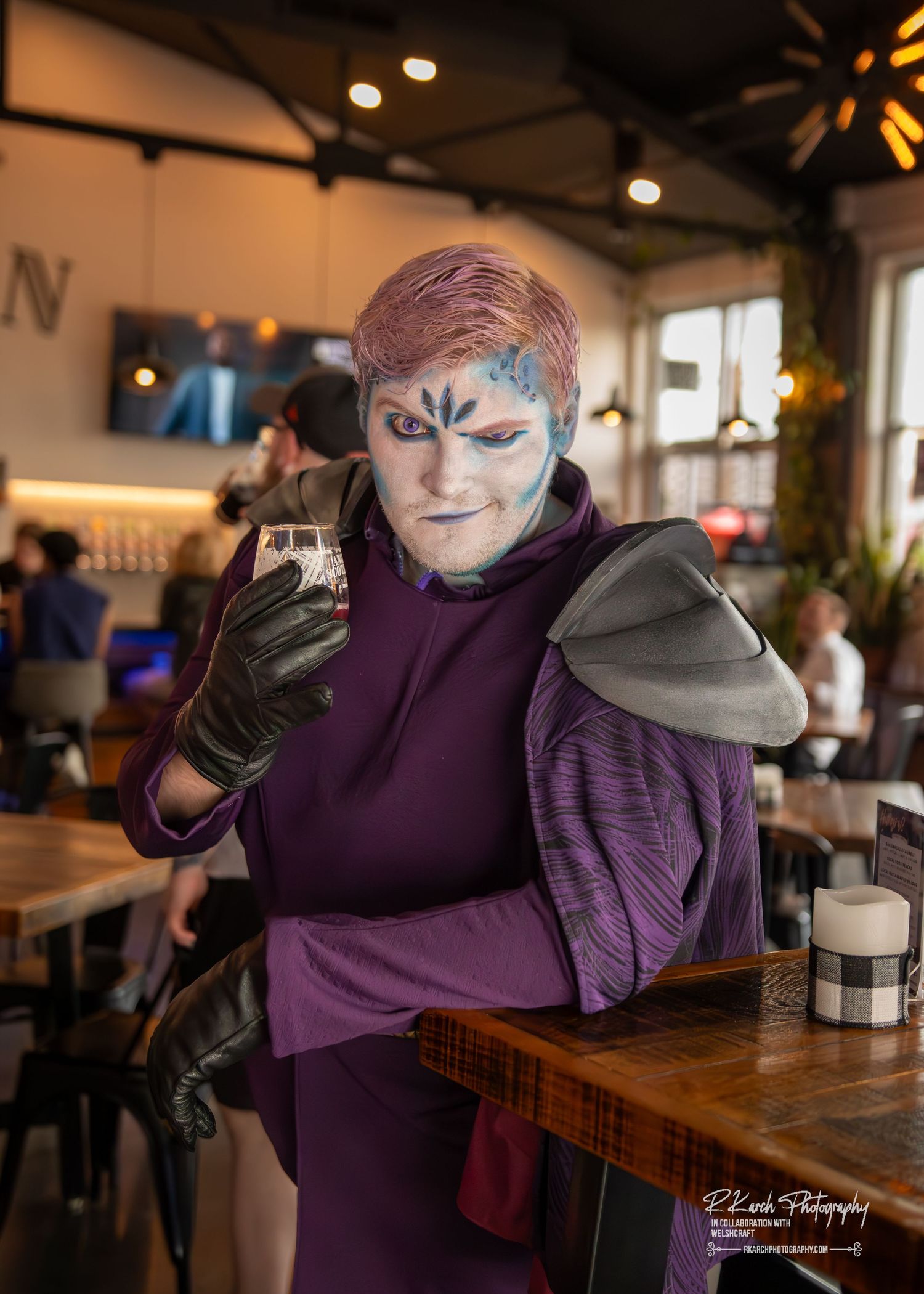 A person in cosplay wearing purple clothing and face paint poses dramatically at a wooden table in a cafe setting.