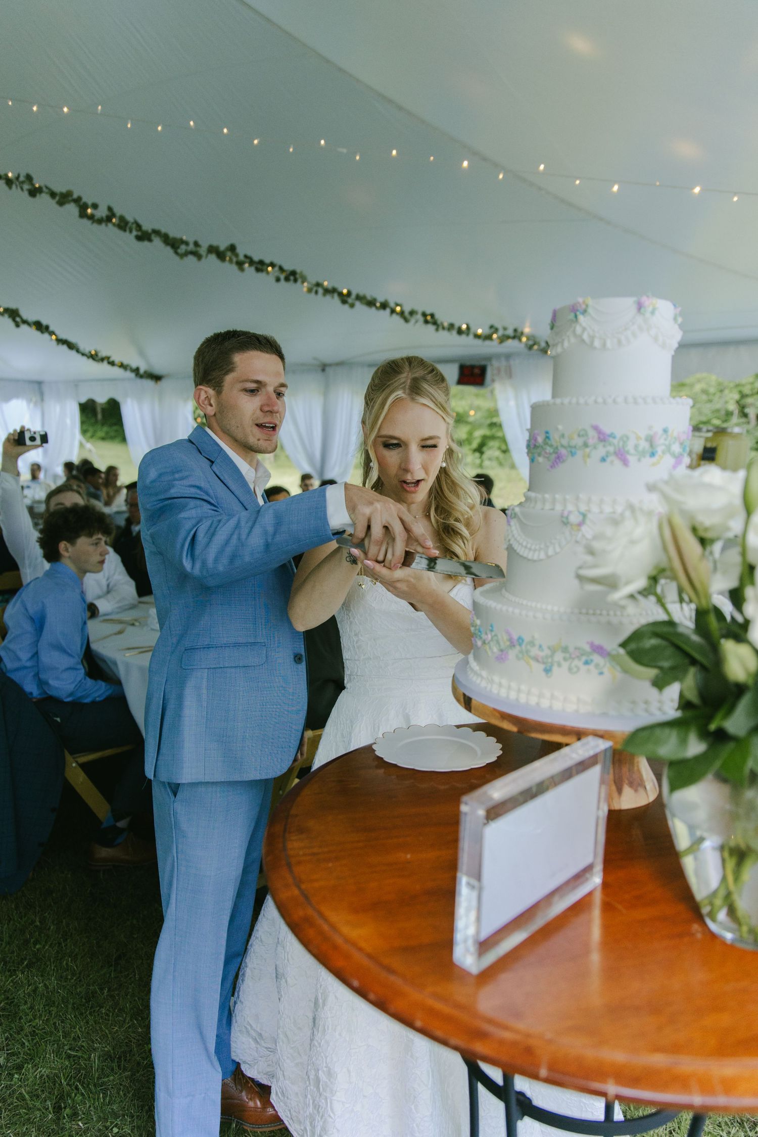 Newlyweds cut their elegant white wedding cake together under a decorated tent.