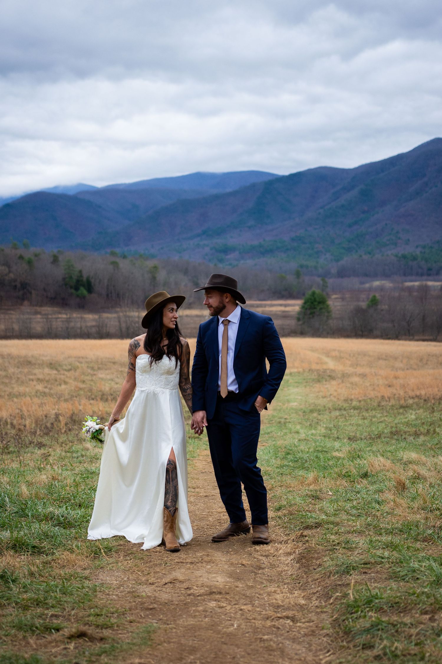A couple in wedding attire walks along a dirt path with rolling mountains and cloudy skies in the background.