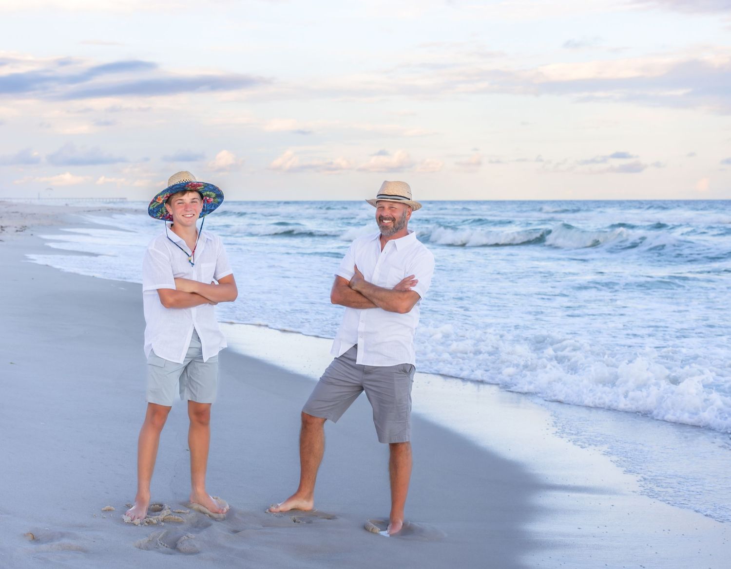 Three people embrace and laugh together on sandy beach at sunset with ocean waves in background.