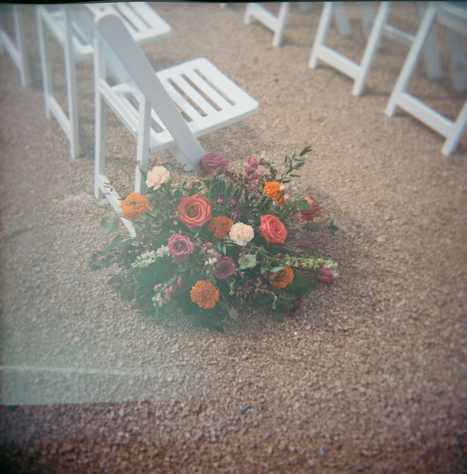 Orange and pink floral arrangement on ground with white folding chairs in background.