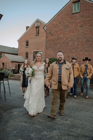 A wedding party in dark tan jackets walks outside a brick church building in a procession line.