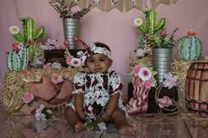 Baby wearing floral romper sits among pink desert-themed decorations with cactus plants and metallic vases.