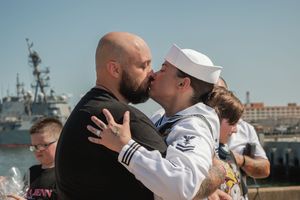 A sailor in white uniform embraces and kisses a loved one during a homecoming at a naval dockyard.