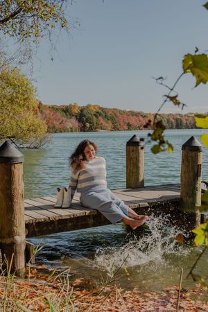 Person in sweater sits on wooden dock splashing feet in lake surrounded by autumn foliage on sunny day.