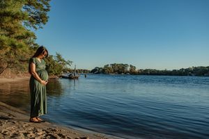 A person in a green dress stands at the edge of a serene lake on a sunny day, surrounded by trees and calm waters.