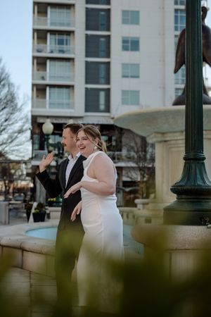 A couple in formal attire share a candid laughing moment next to a lamp post in an urban setting with a high-rise building behind them.