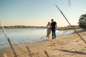 Two figures walk hand in hand along a peaceful beach at sunset with coastal waters and trees in view.