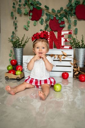 First birthday photo setup with red and green decor featuring a small child sitting among apples and wooden crates.
