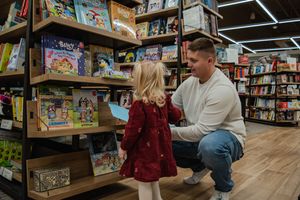 Two people browse children's books together at a bookstore with wooden floors and shelves full of colorful titles.