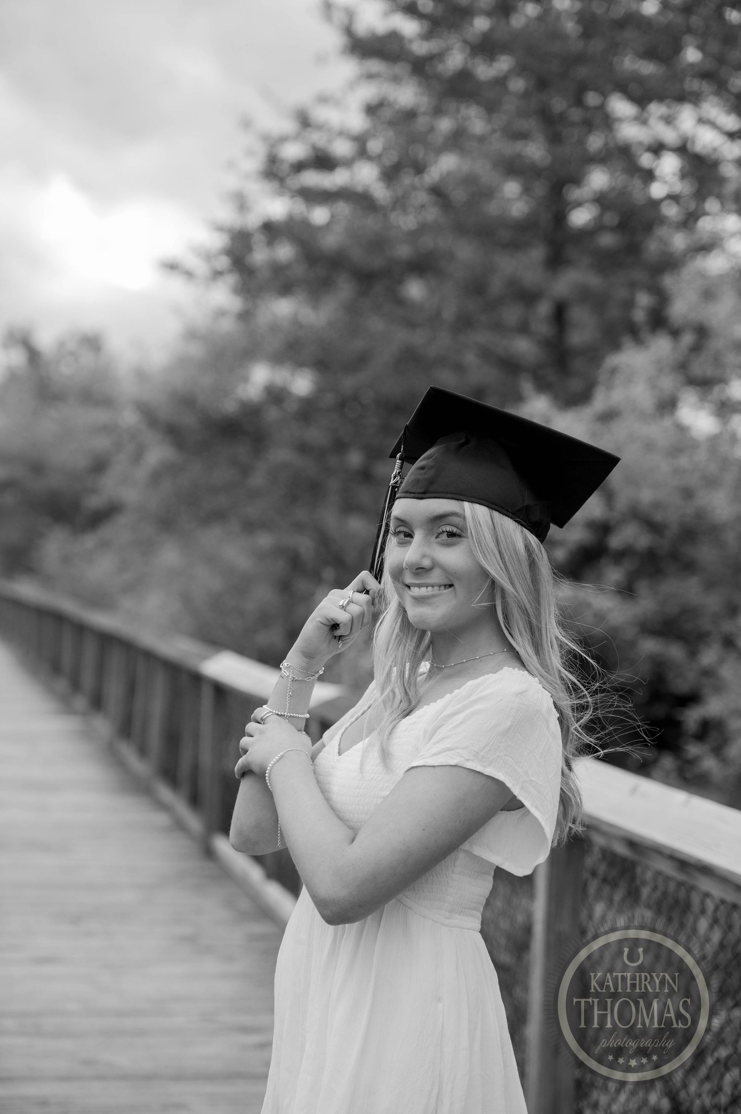 Black and white graduation photo showing a graduate in cap and gown smiling on a wooden bridge.