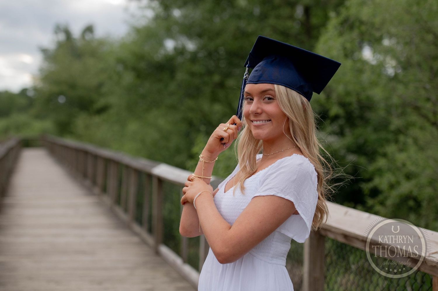 A graduate in white t-shirt and cap stands smiling playfully on a wooden bridge among trees.