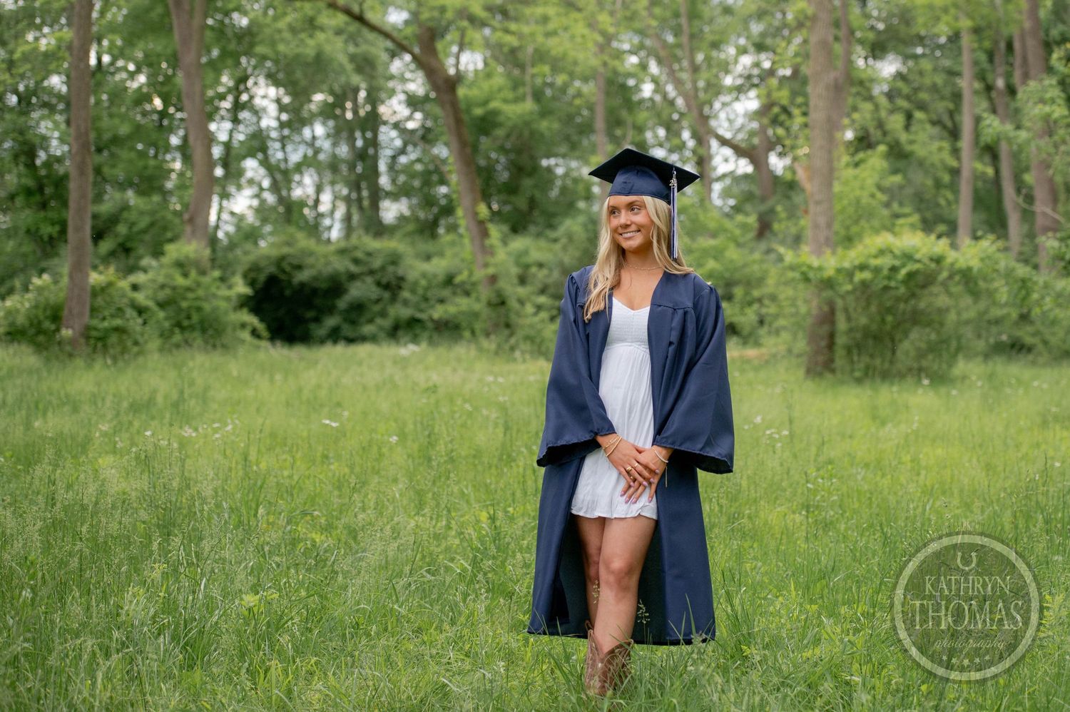 Graduate in blue cap and gown stands in grass field with trees in background during outdoor graduation photo session.