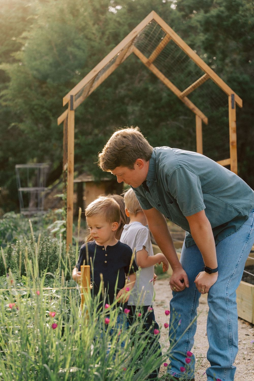 Two people examining plants and flowers in a garden under a wooden arch structure on a sunny day.