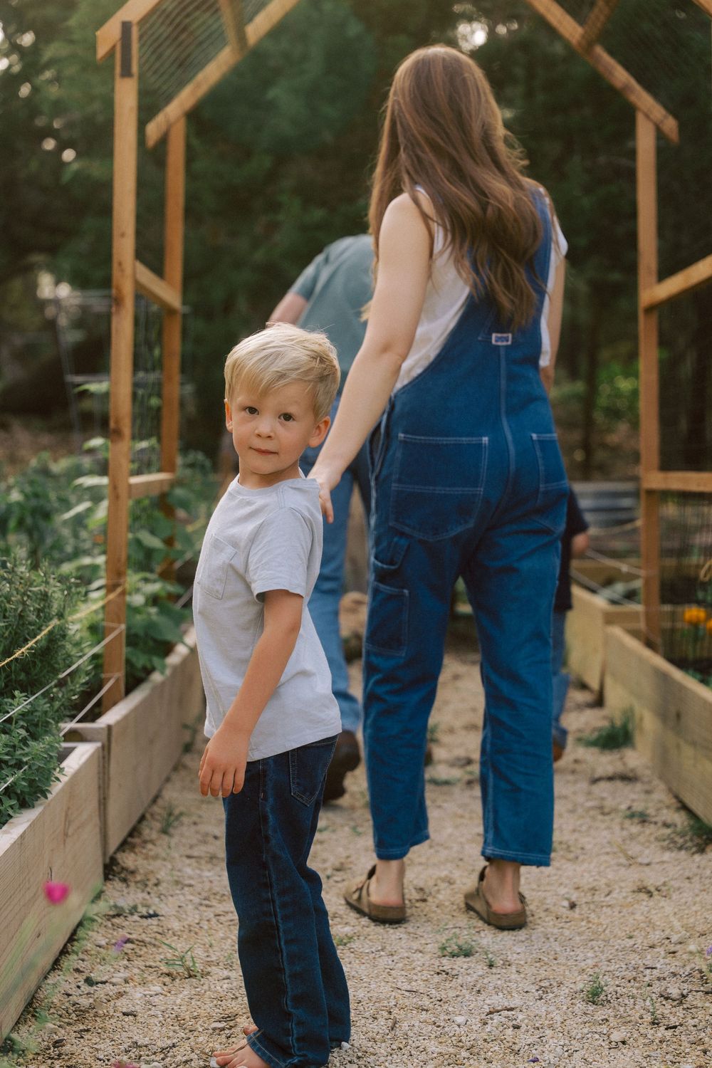 Two people standing in a garden pathway next to raised wooden planter beds under a wooden arbor.