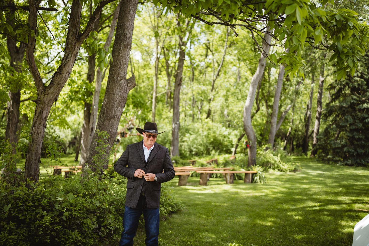 A figure stands in a lush garden setting surrounded by tall trees and green grass on a sunny day.