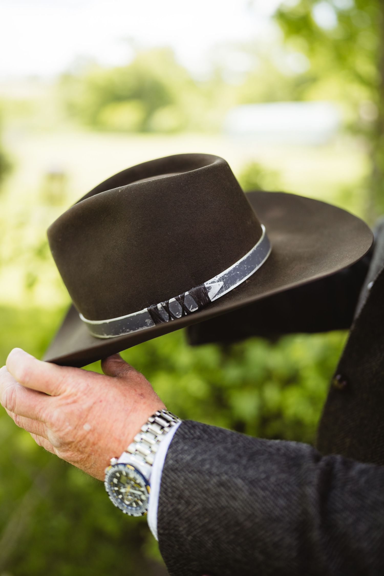 Brown fedora hat with silver band held up against blurred green outdoor background while wearing silver watch.