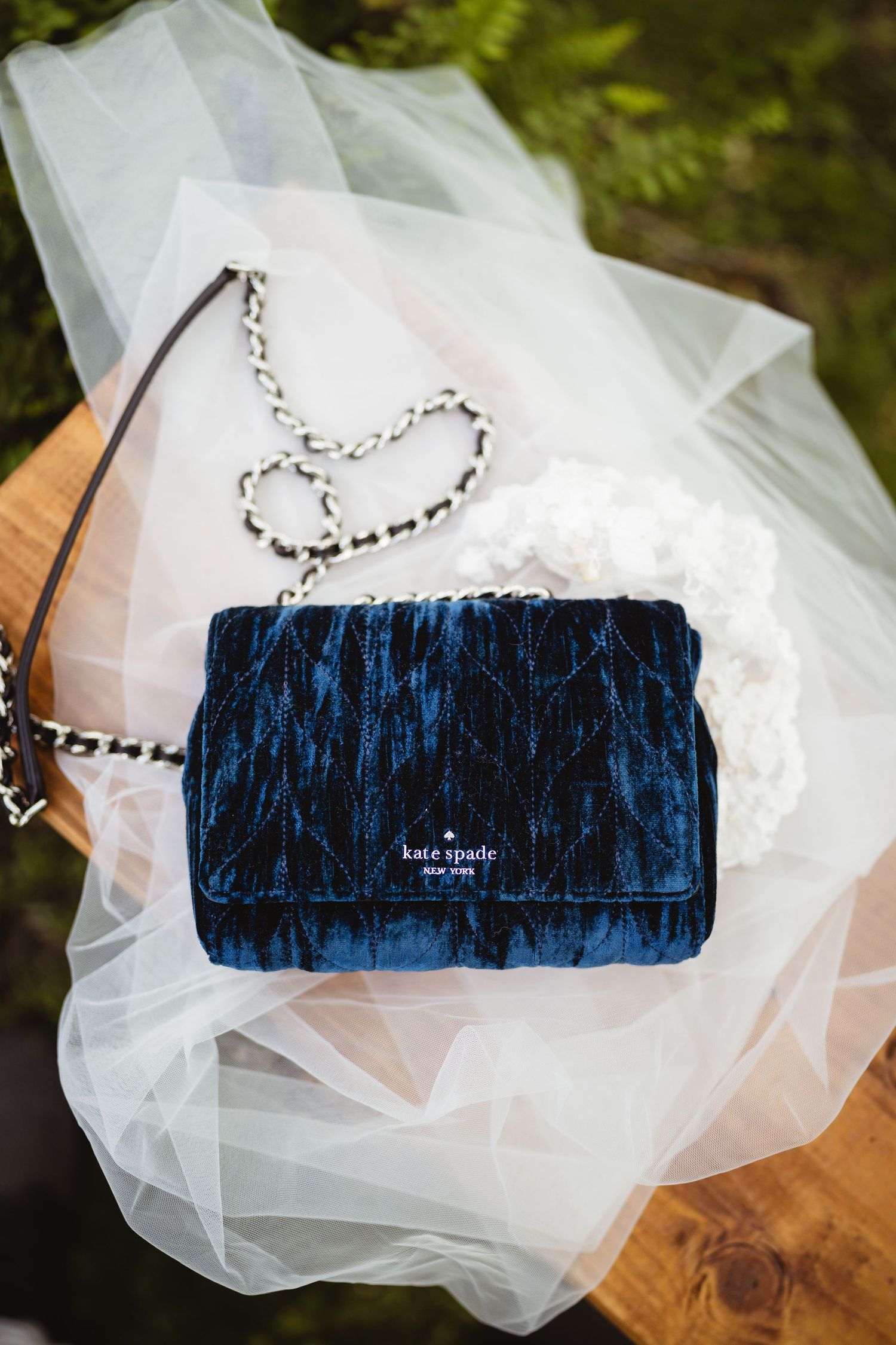 Elegant blue velvet Kate Spade handbag with silver chain strap displayed on white tulle fabric and wooden surface.