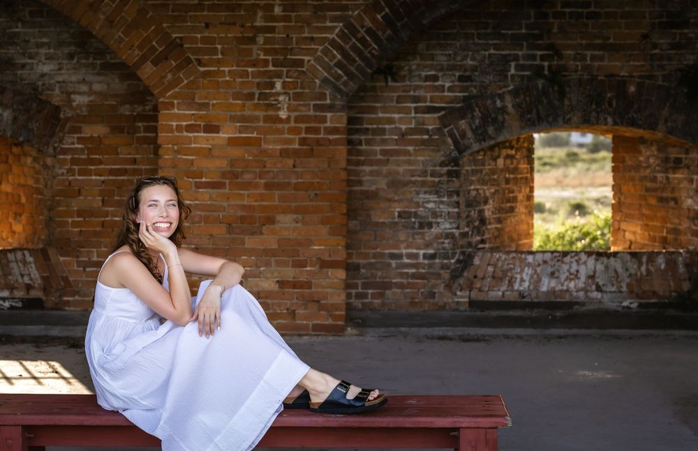 Person in white dress sits on red bench inside brick archway structure with window opening to nature view.