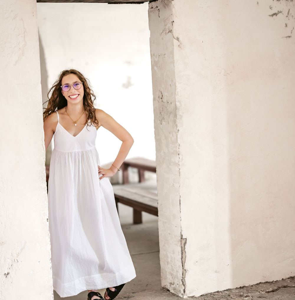 A person in a flowing white sundress stands smiling in a rustic white archway.