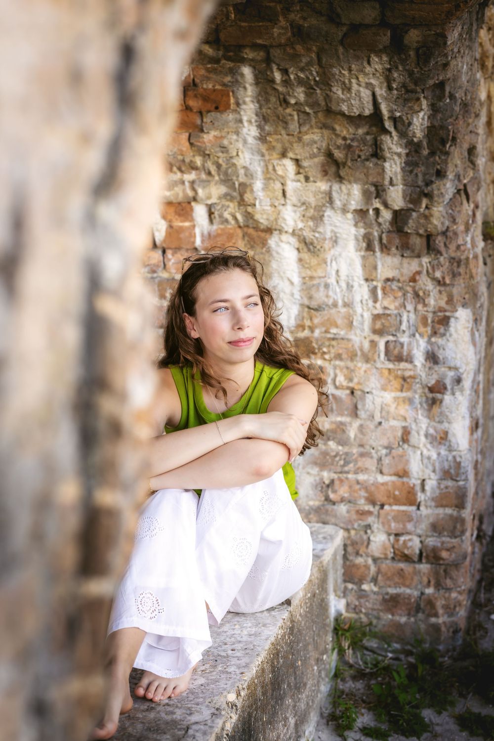Someone in a lime green top sits thoughtfully in an old brick archway while wearing flowing white pants.