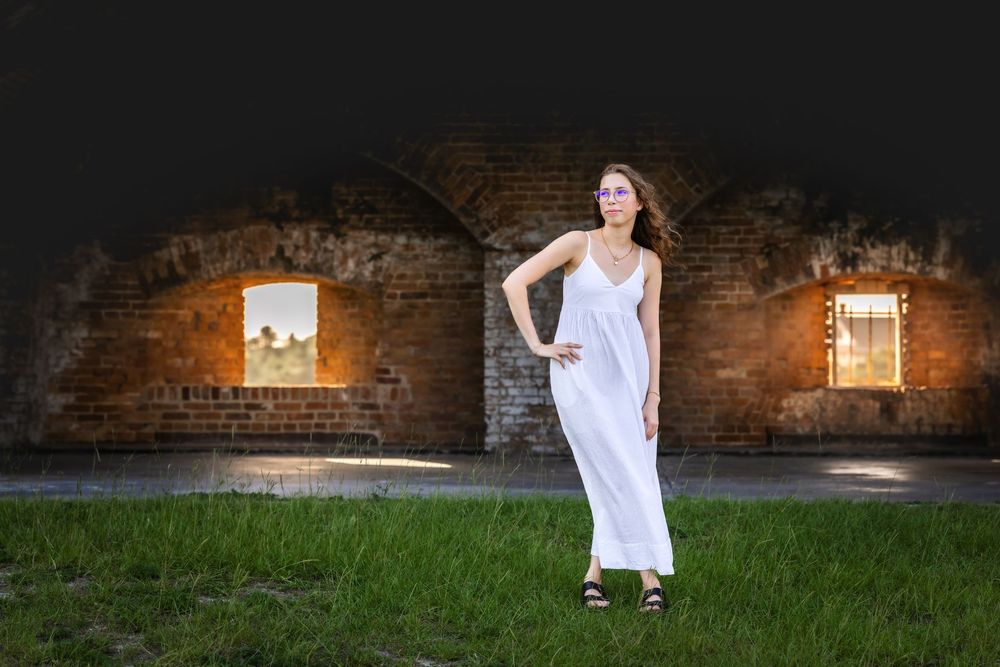 Person in flowing white dress poses elegantly against rustic brick wall backdrop with dramatic lighting and shadows.