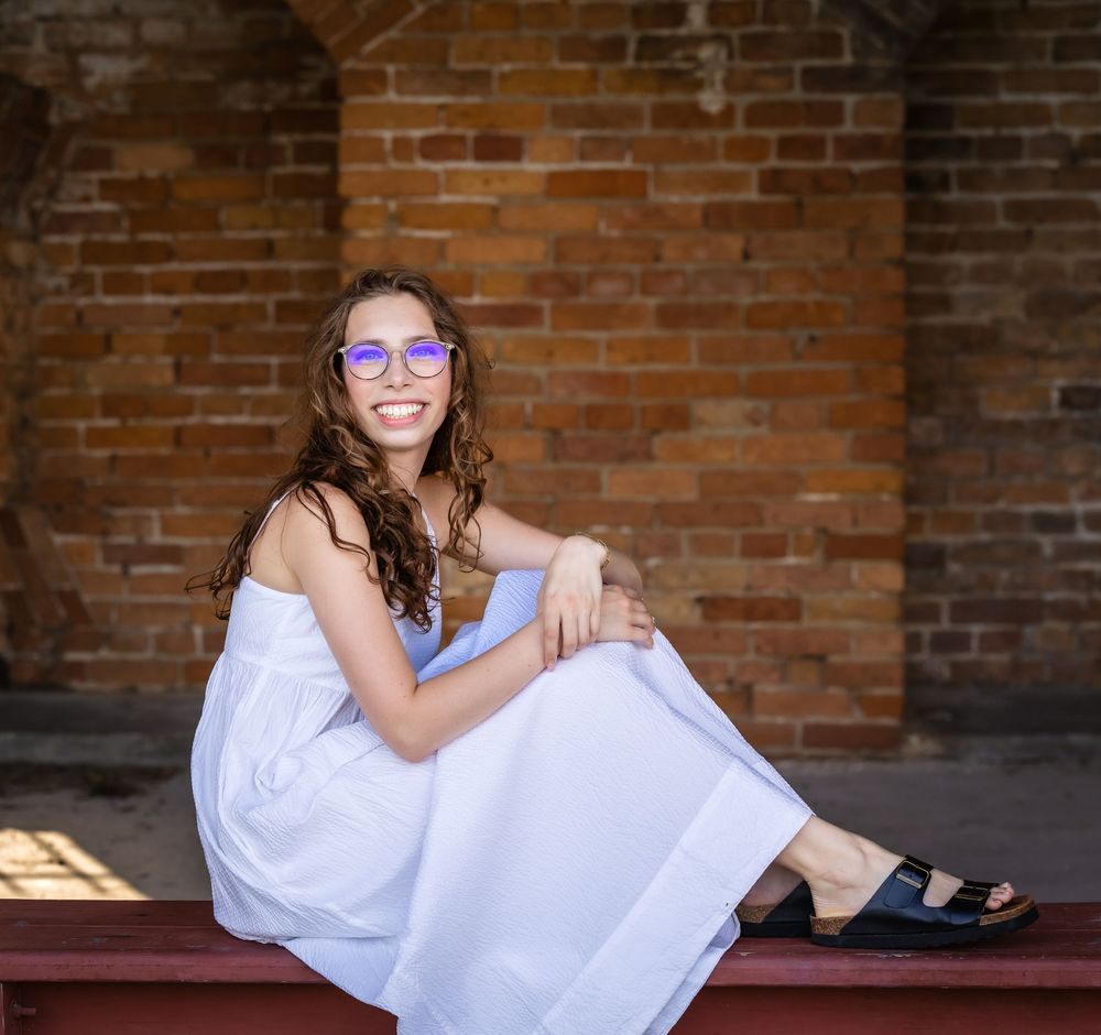 A relaxed figure in a white dress sits on wooden steps against a rustic brick wall with dramatic lighting.