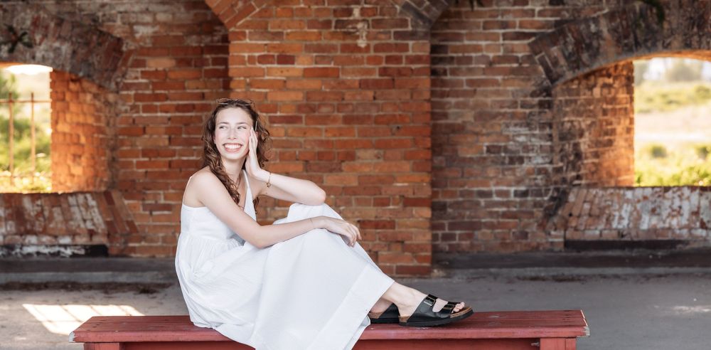 Person in a white dress sits on a red bench against an exposed brick archway in warm sunlight.