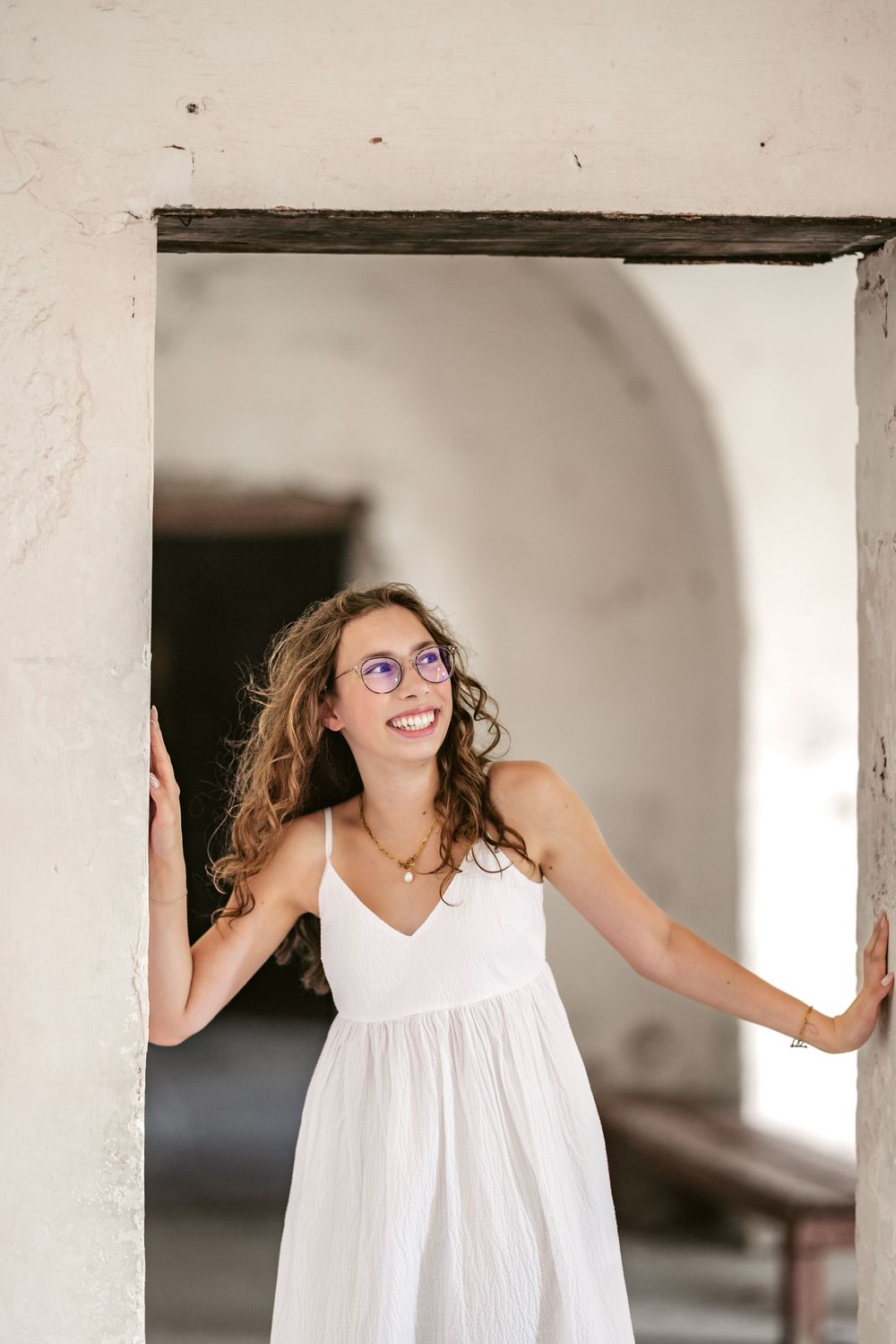 A person in a white sleeveless dress poses playfully between architectural white columns.