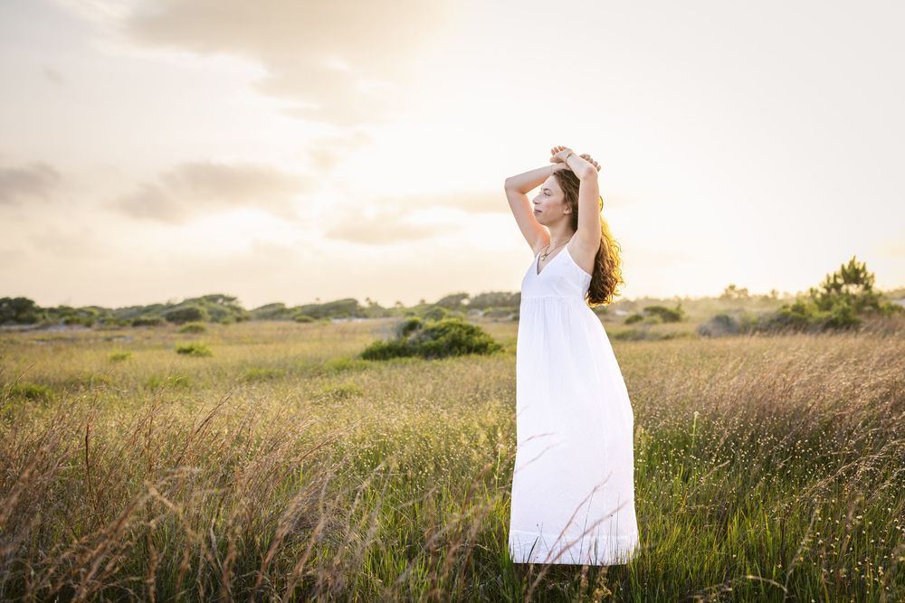 A person in a long white dress poses in a golden field at sunset.
