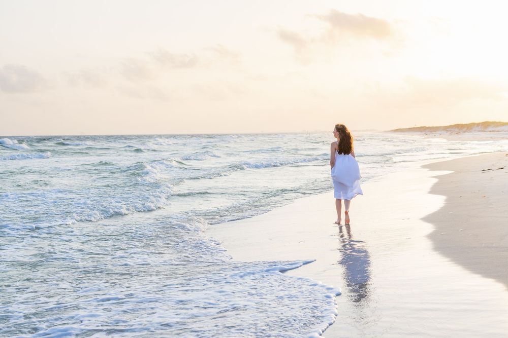 A figure in a light dress walks alone along a serene beach shoreline during sunset as waves roll in.