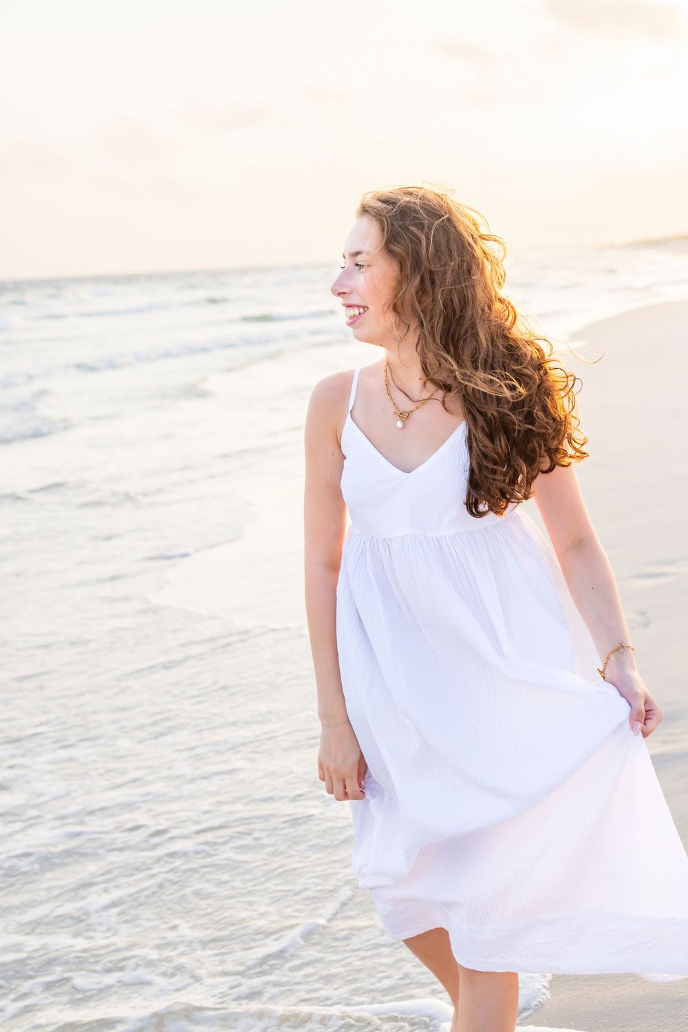 Someone in a flowing white sundress enjoys a carefree moment on the beach at sunset while their hair blows in the breeze.