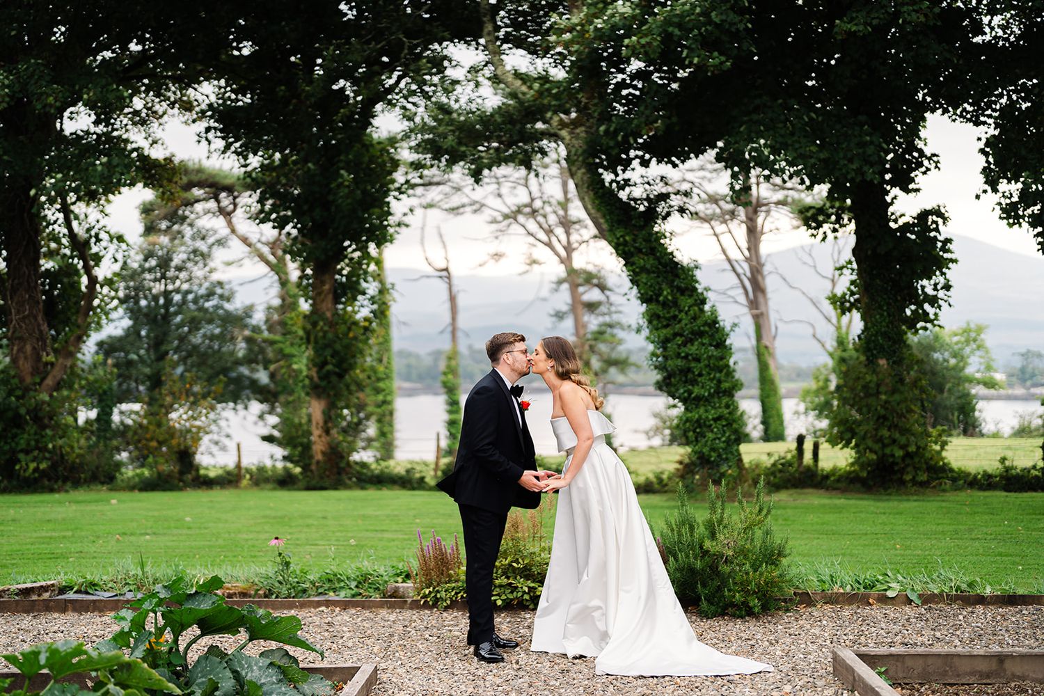 A romantic wedding portrait of a couple standing among trees with a lake view.