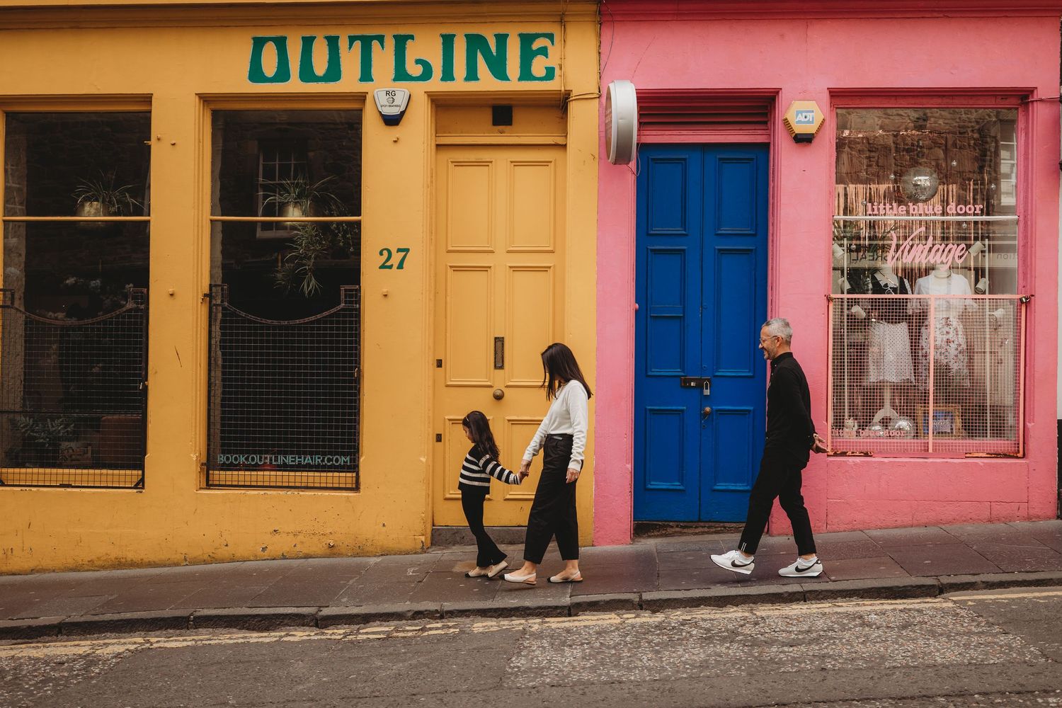 People walking past colorful storefronts with yellow and pink facades and a blue door on a charming city street.