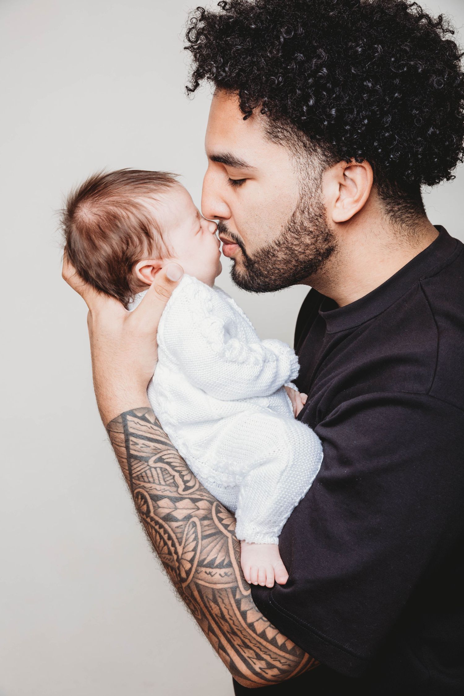 A person wearing a black sweater with a tribal tattoo sleeve holds and kisses a baby dressed in white.