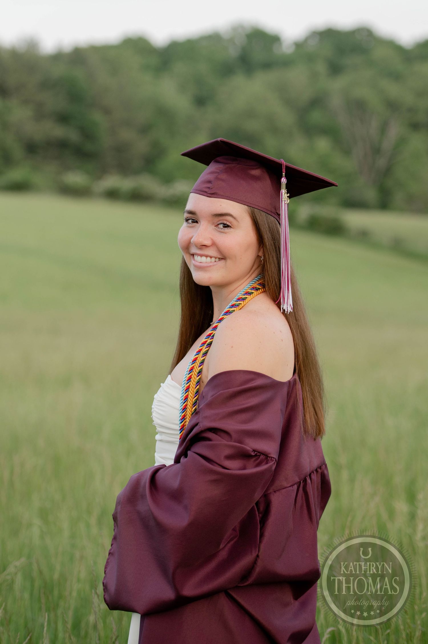 Graduate in maroon cap and gown poses in a grassy field during an outdoor graduation photoshoot.