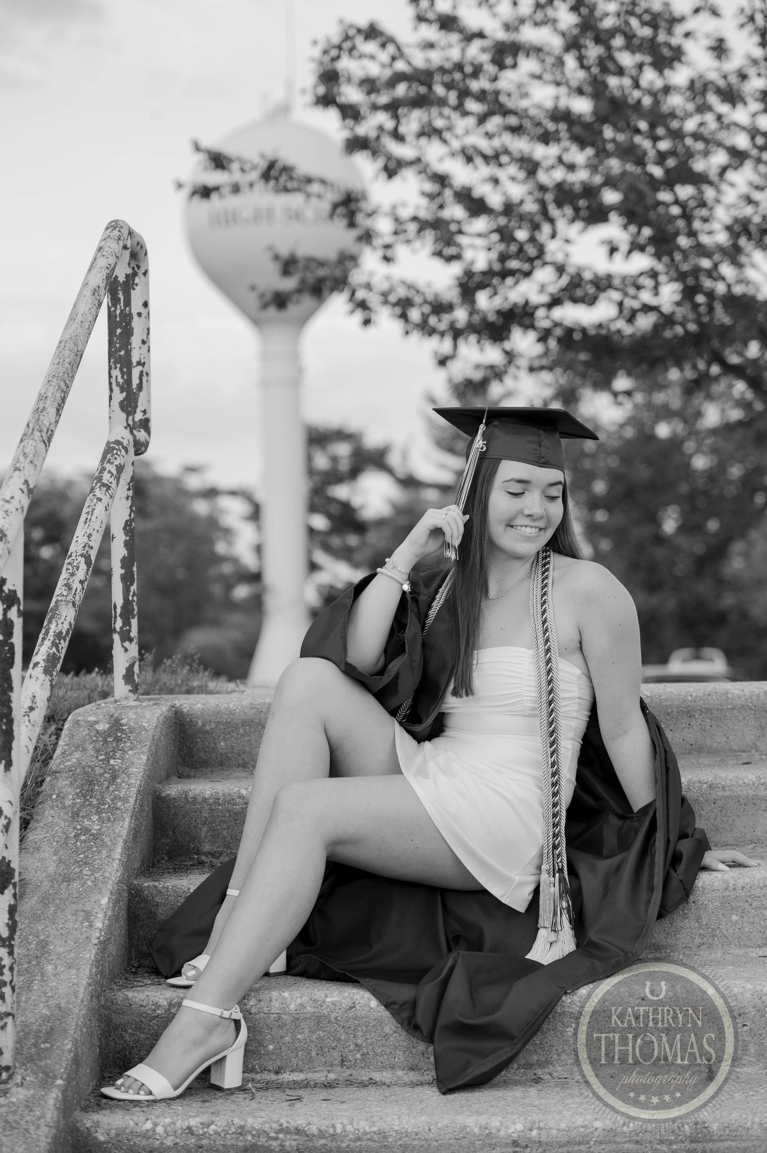 Black and white graduation portrait of a graduate sitting on steps with a water tower visible in the background.