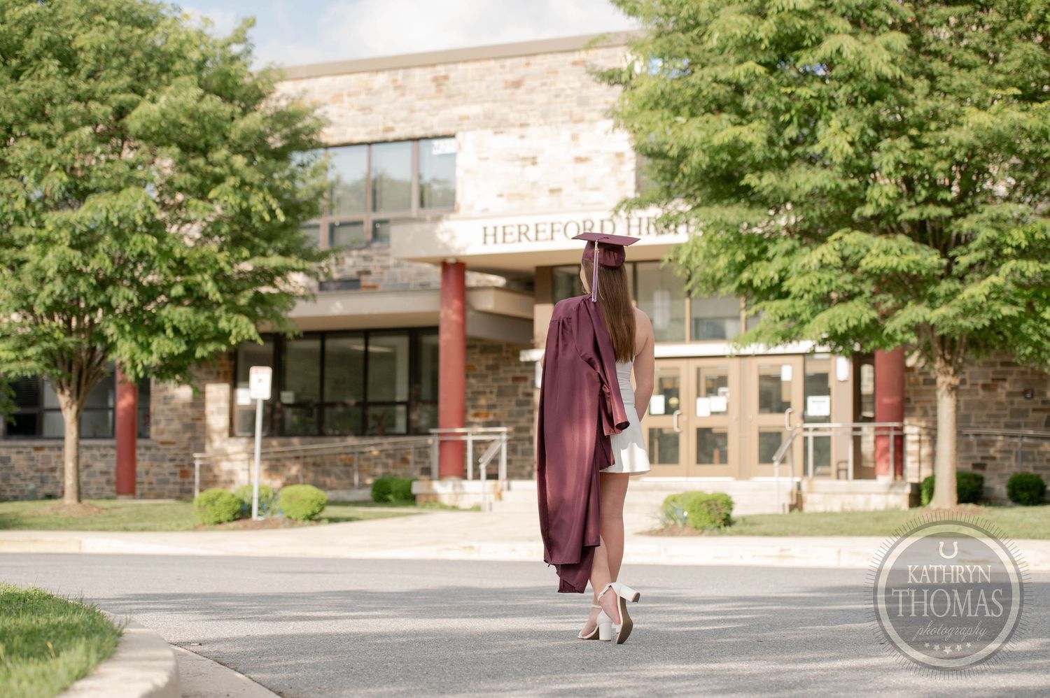 Graduate in burgundy cap and gown poses in front of brick university building entrance surrounded by green trees.