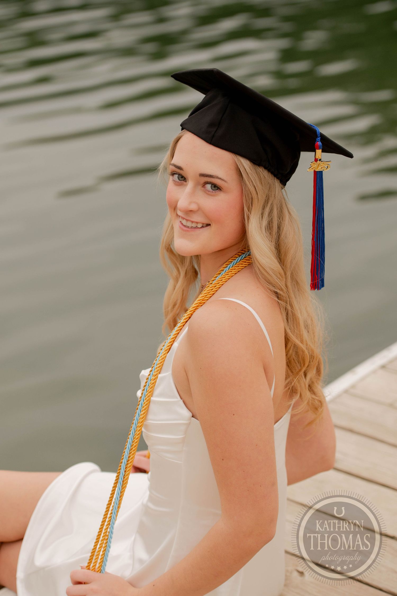 Graduate in white dress and cap sits on dock by water with honors cords and tassel on a sunny day.