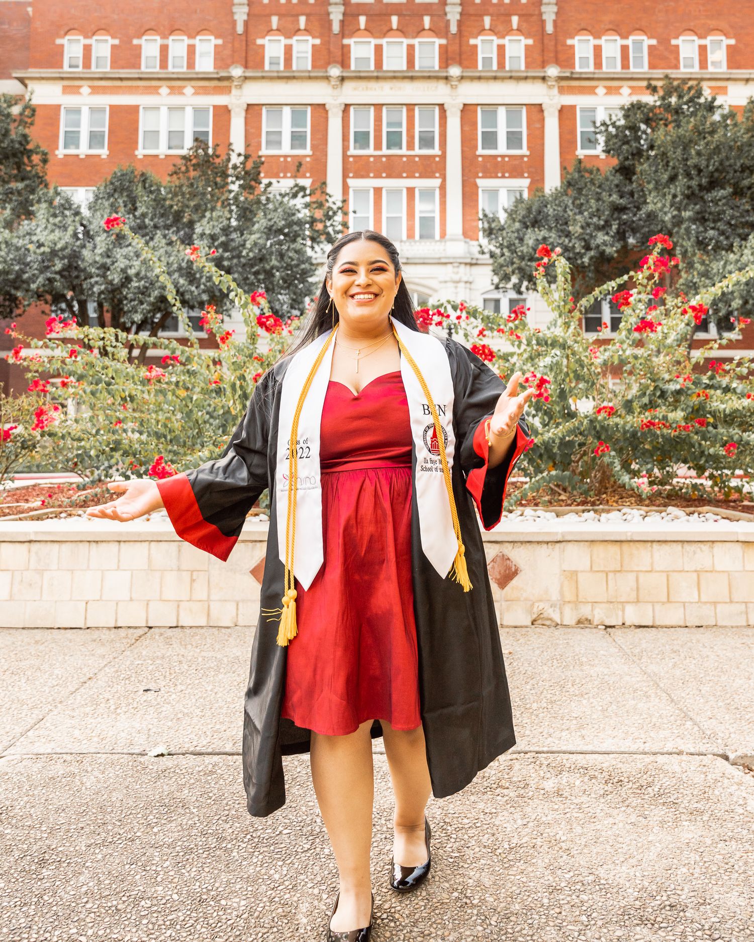 Graduate wearing red dress and graduation regalia poses joyfully in front of brick university building.