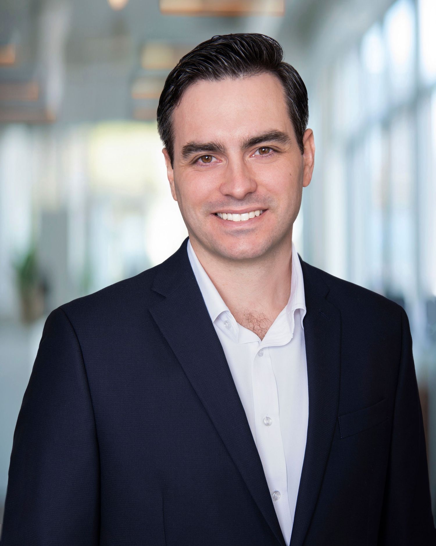 Professional headshot of a person in a dark suit and white button-down shirt against a bright office background.