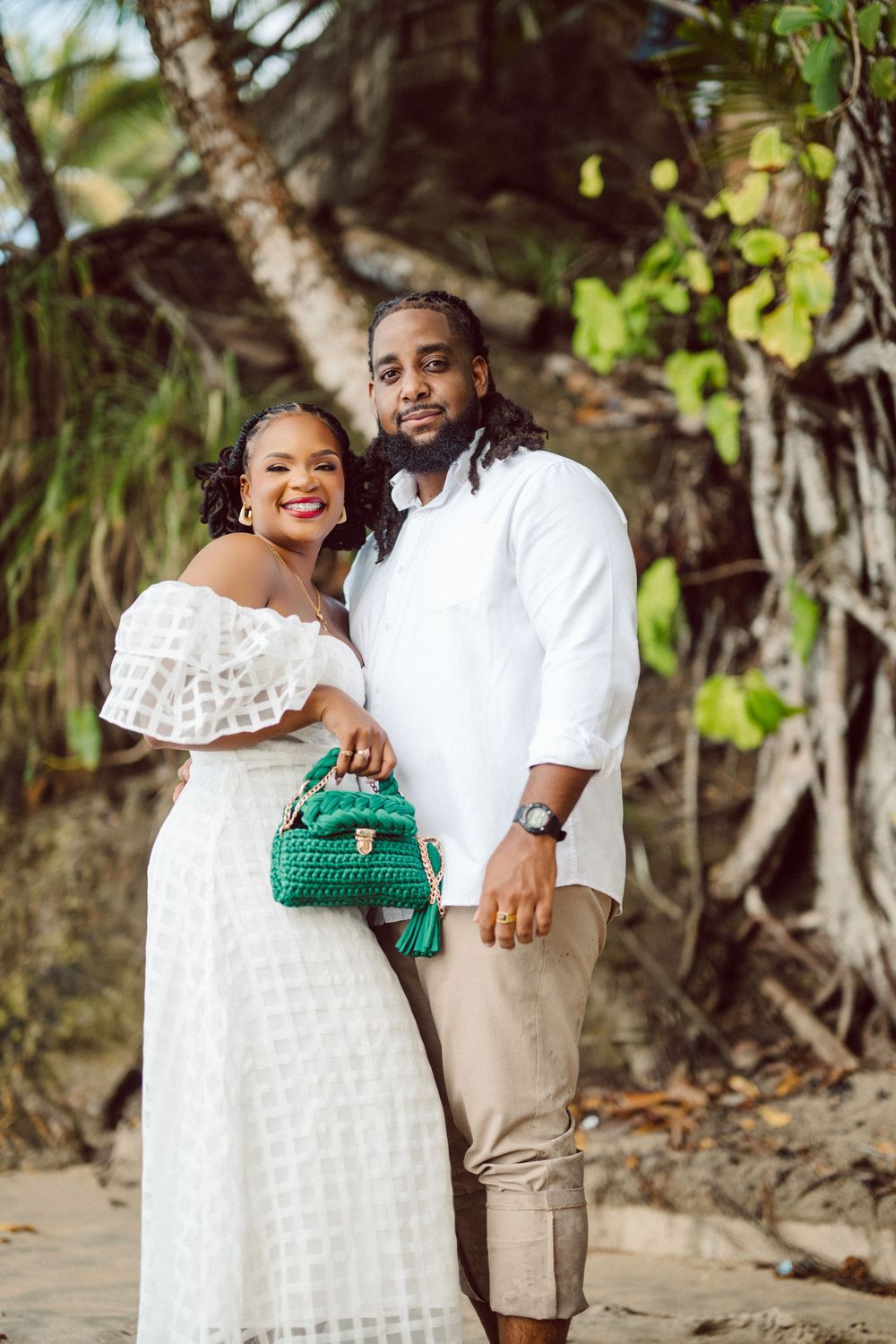 A couple poses on a beach wearing coordinated white outfits with the woman holding a green handbag near tropical trees.