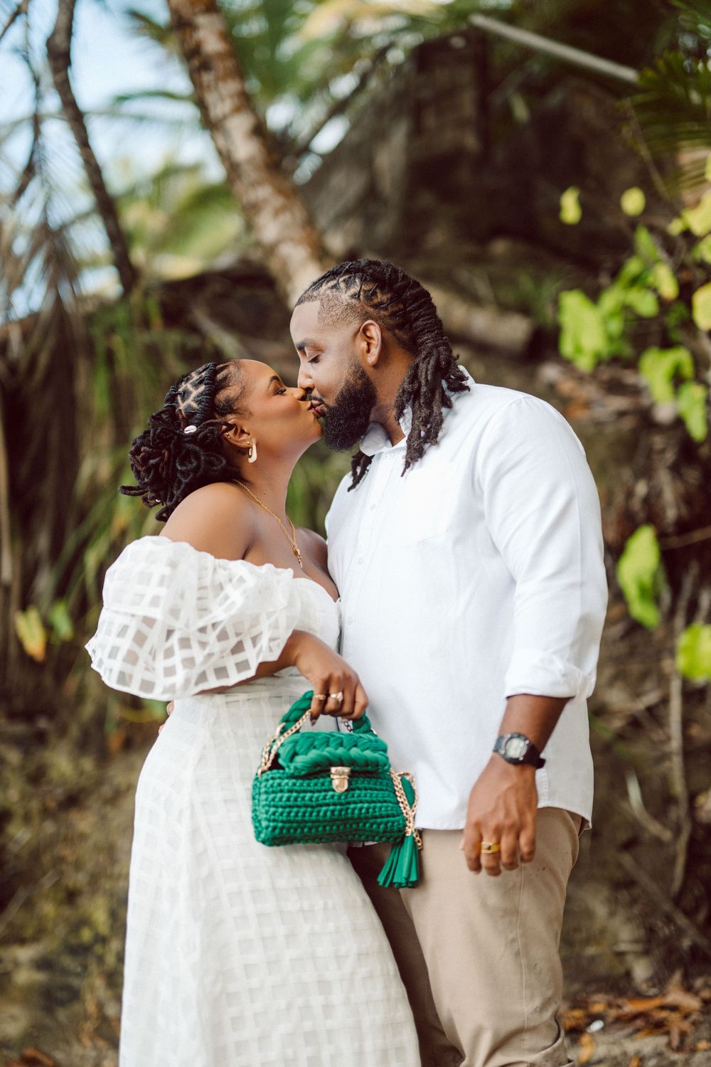 Romantic couple in coordinated white outfits share a tender moment outdoors while holding a green woven handbag.
