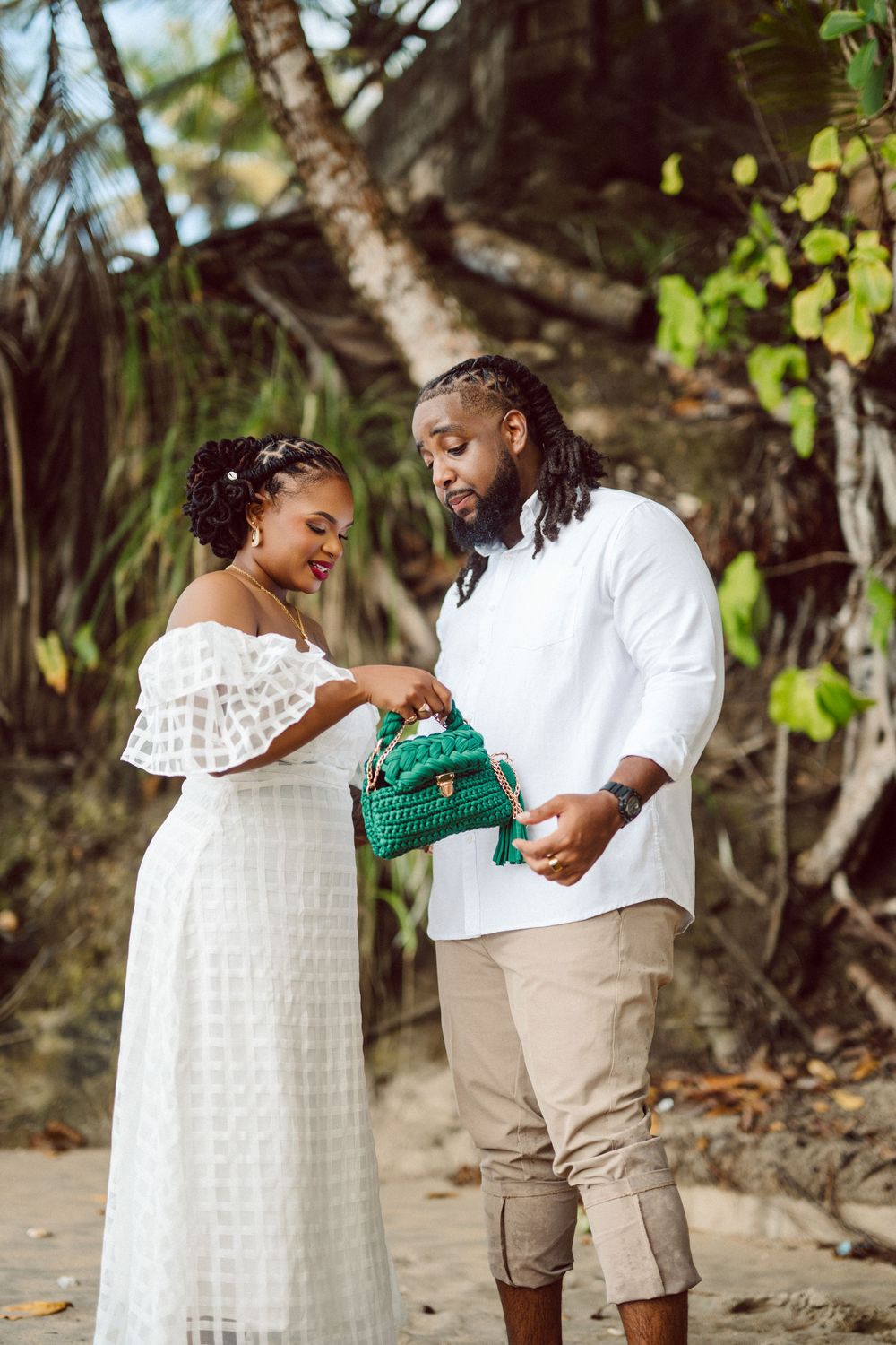 A couple in white outfits sharing a moment on the beach while looking at an emerald green handbag.