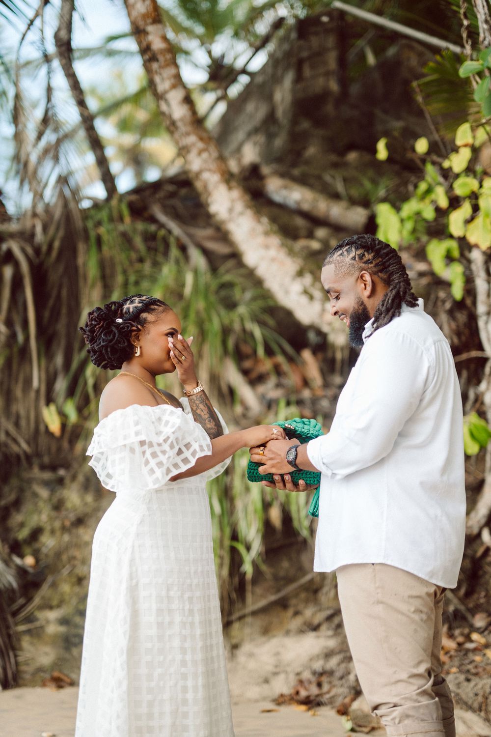 A romantic beach wedding moment shows a couple in white attire exchanging rings among palm trees and tropical foliage.