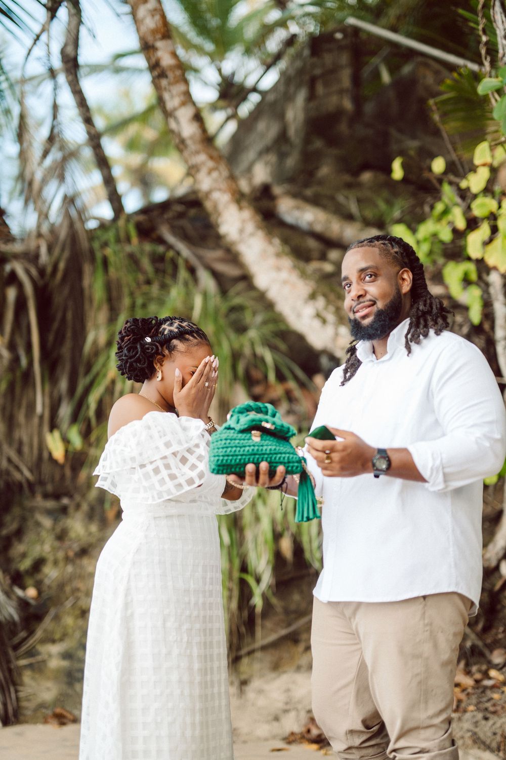 A couple shares a joyful moment on the beach as one presents a wrapped gift to the other who covers their face with excitement.