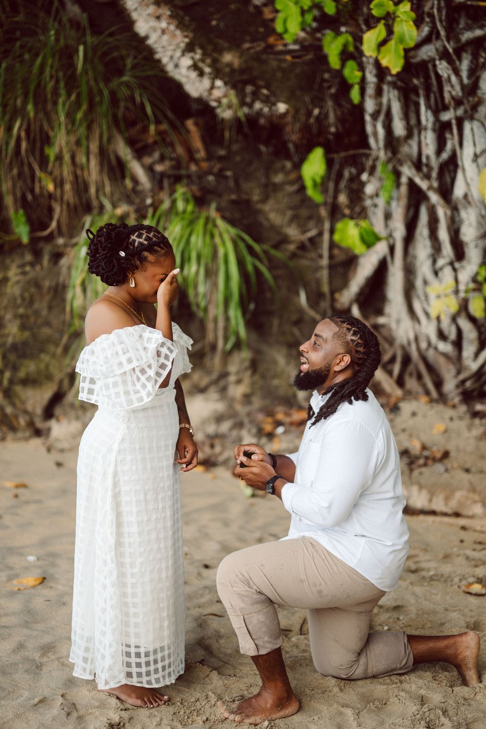 A romantic beach proposal scene with two people in white outfits on the sand with tropical foliage in the background.