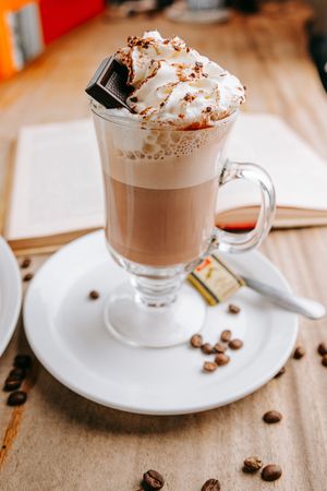 Four glass mugs of hot chocolate topped with whipped cream and cocoa powder on a wooden table with scattered coffee beans.