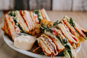 Club sandwich with layers of meat, lettuce and tomato served on a white plate with creamy dipping sauce.
