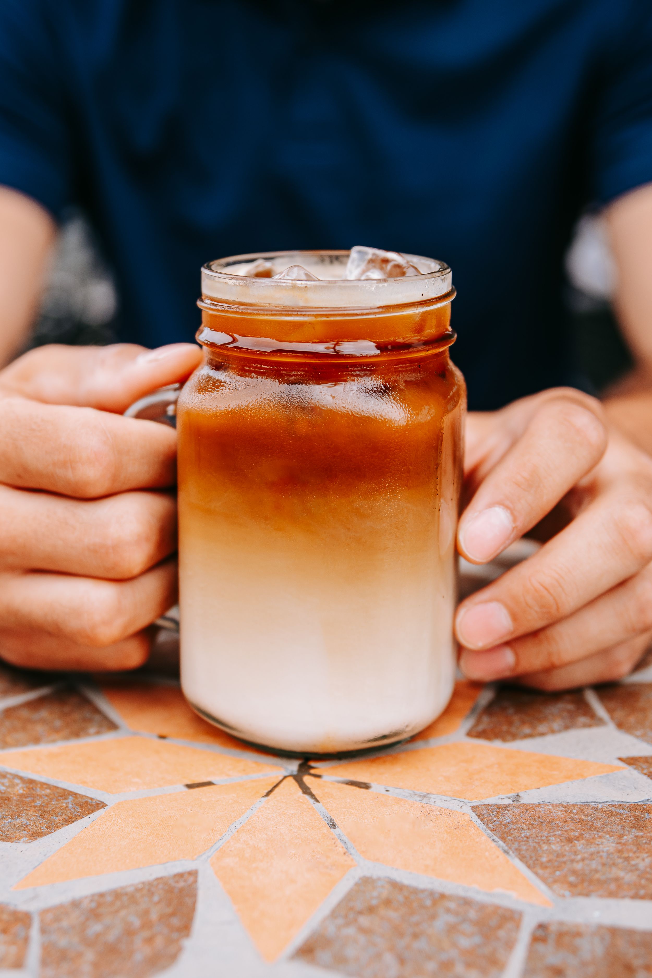 A sequence of hands holding ombre-colored iced coffee drinks in mason jars on a patterned surface.
